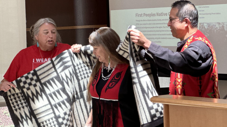 A man and a woman place a decorative blanket around another woman