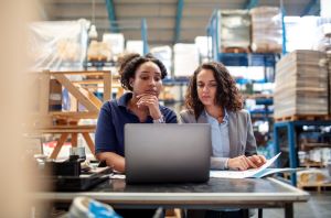Two women sit at a desk looking at same laptop