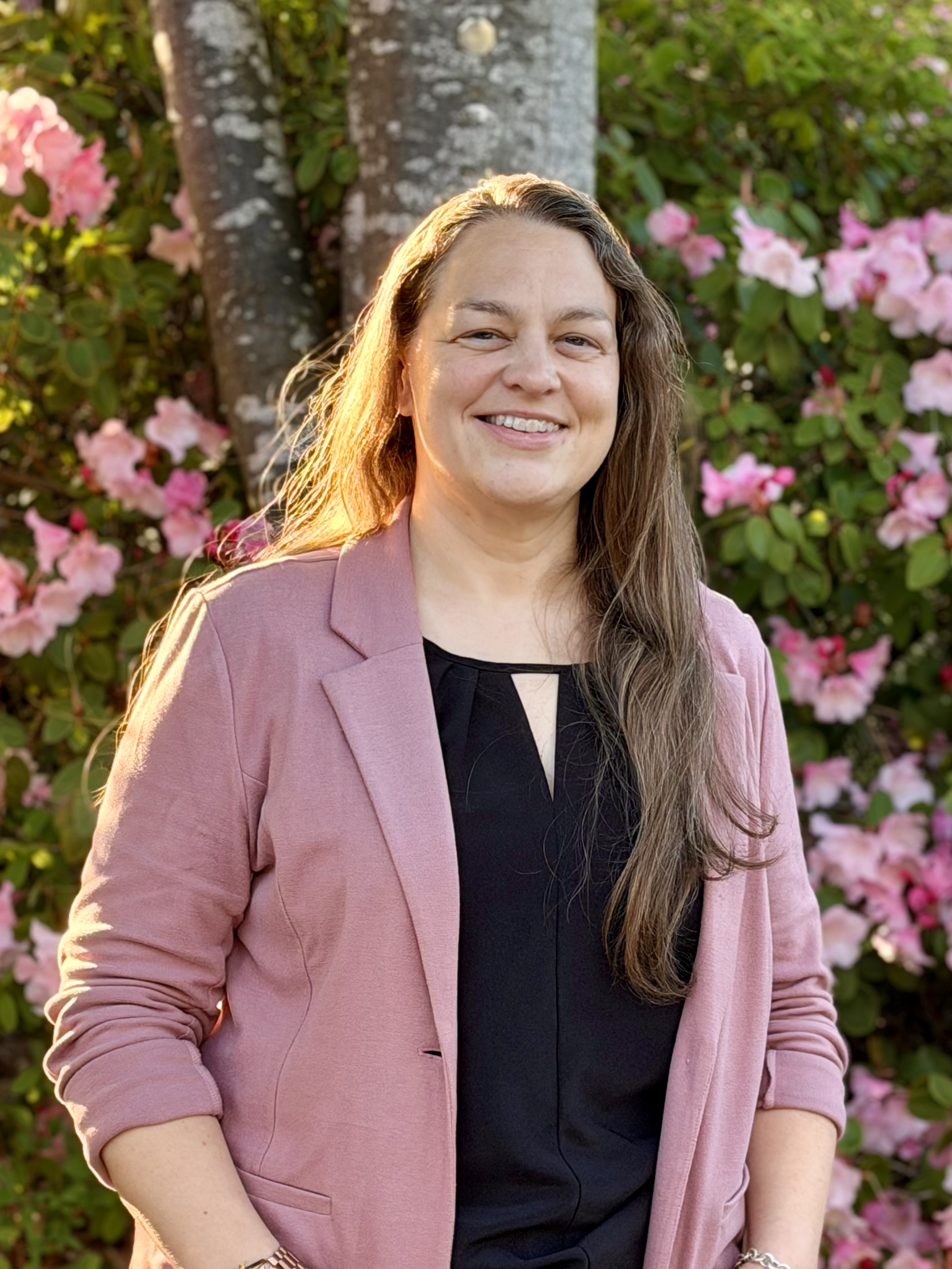 Chara Powell wearing a black blouse and dusty rose suit jacket standing in front of trees and flowers
